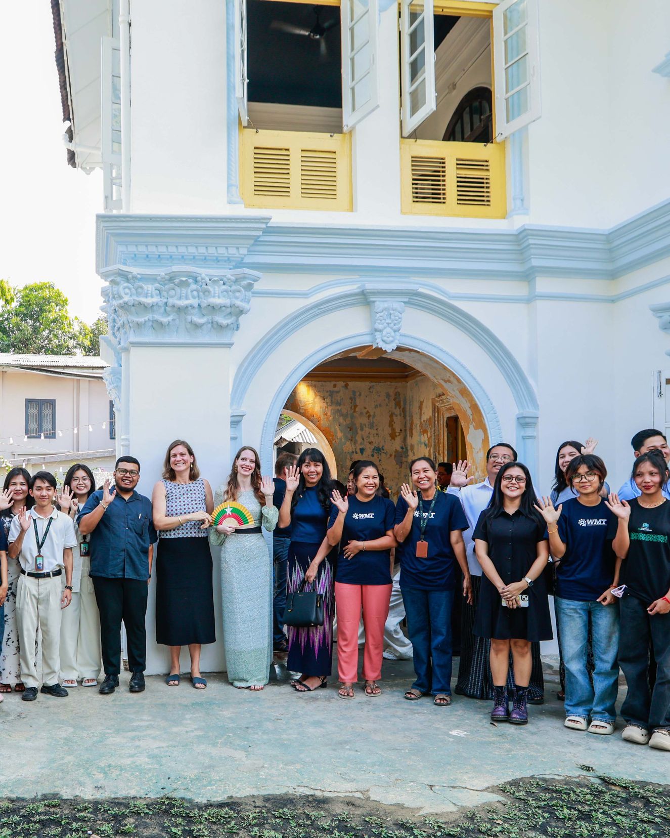 Women in team T-shirts and guests wave in front of the restored white facade and arched entrance of the Thanlyin HERitage Centre