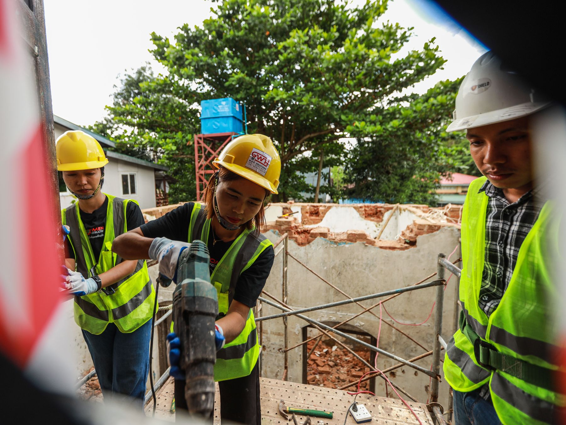 Two women trainees in yellow hard hats and high-visibility vests drill on scaffolding at a building site while a colleague in a white hard hat looks on