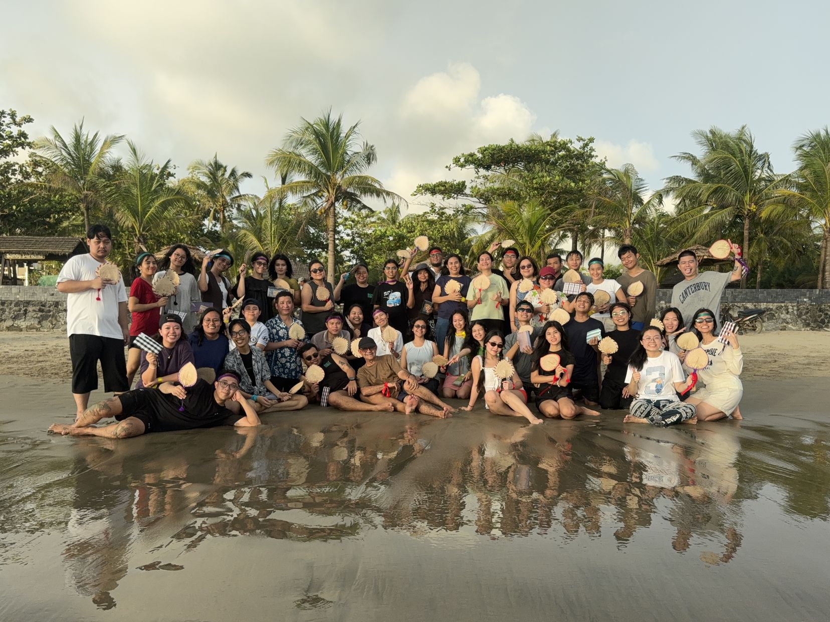 The Doh Eain team gathered for a group photo on a beach at low tide, with palm trees behind and their reflection in the wet sand
